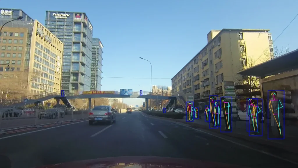 Dash-cam view of a multi-lane urban road in Beijing with tall buildings and a pedestrian bridge, where a group of pedestrians on the right sidewalk are marked with blue bounding boxes and colored skeleton lines.